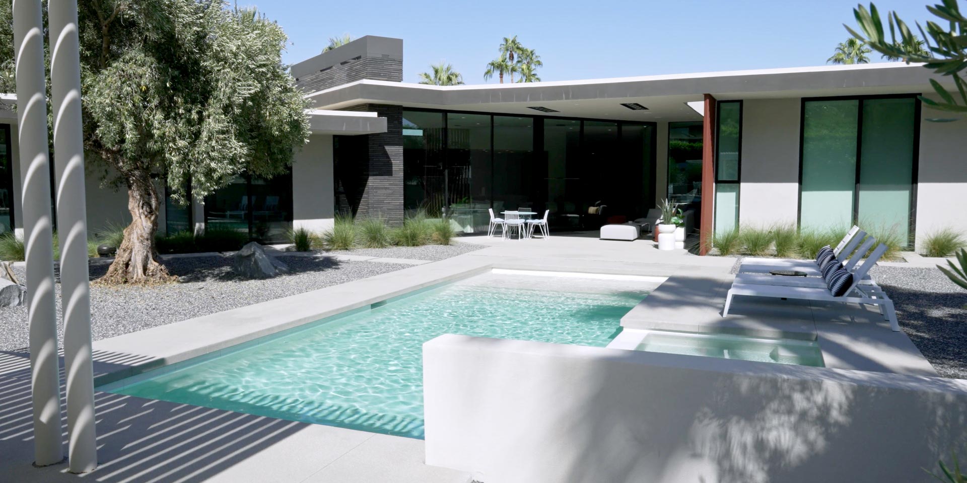 A modern backyard with a rectangular pool and spa surrounded by a green artificial turf lawn, with a hillside and trees in the background.