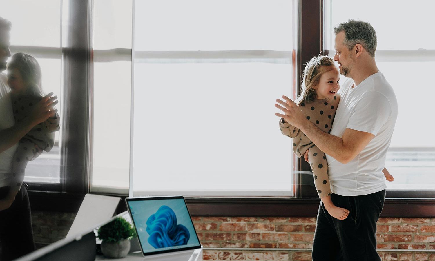 A man holding his daughter in a well-lit room with a brick wall and a laptop displaying a blue abstract image on the desk.