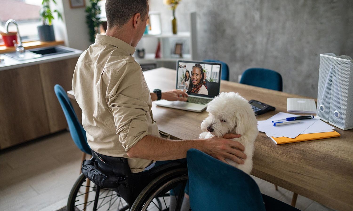 A man in a wheelchair having a video call at a kitchen table, with a small dog sitting next to him.