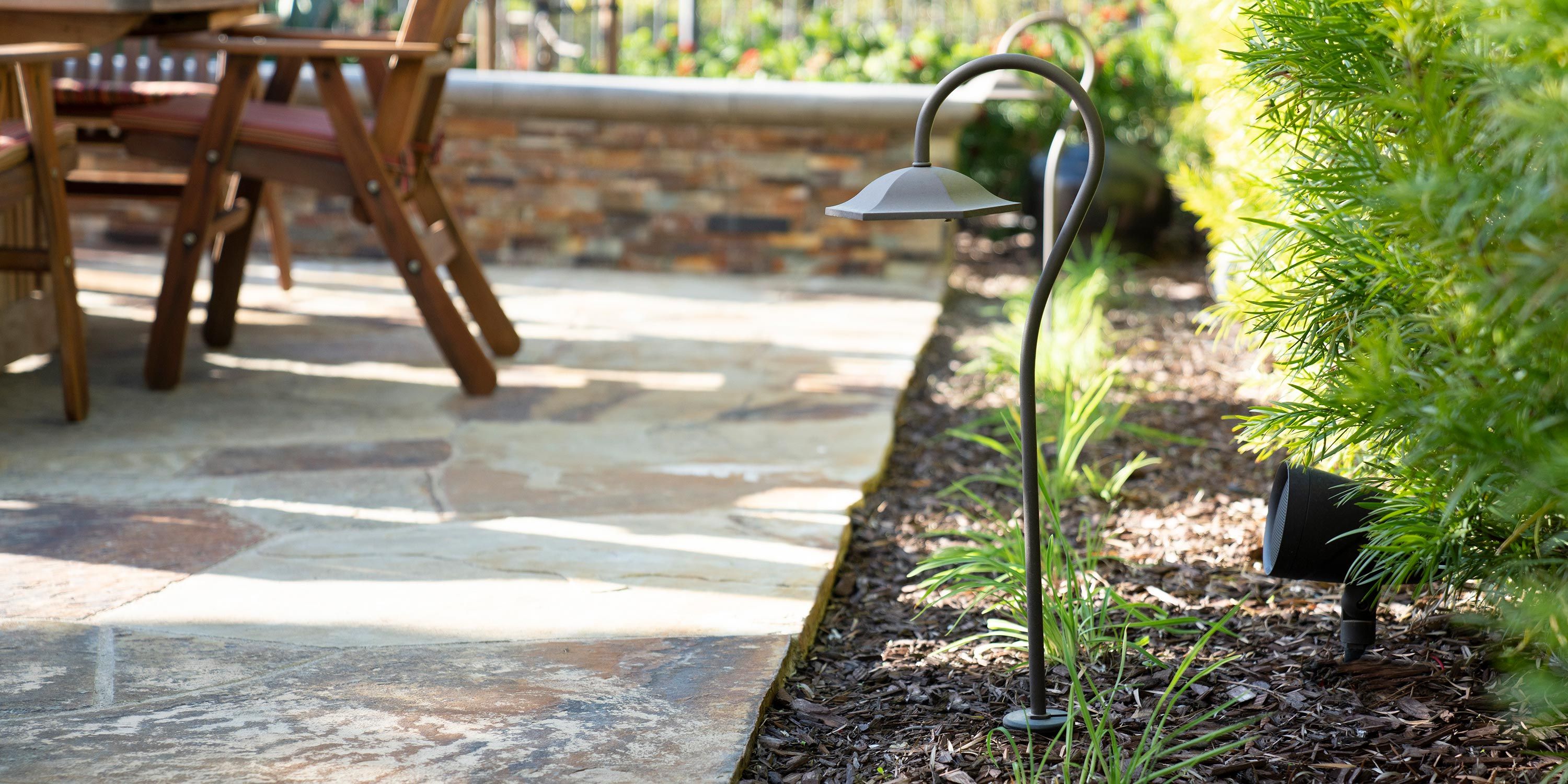 A close-up of a stylish outdoor garden light fixture next to a patio area with wooden chairs in the background.